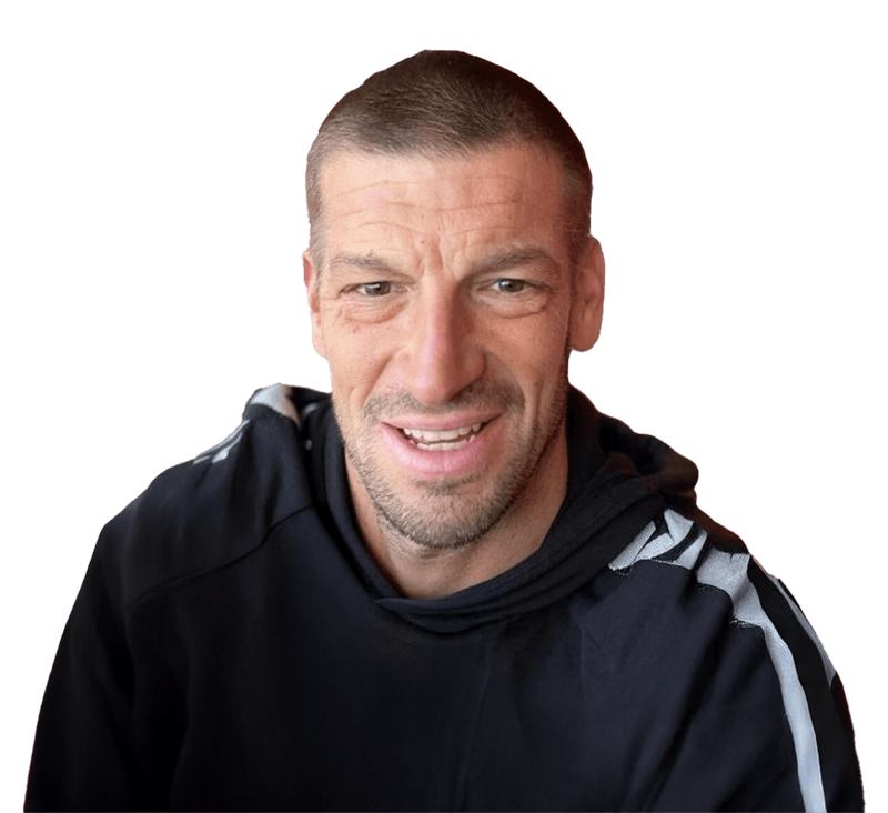 A man with short hair and a trimmed beard smiles whilst wearing a black hoodie with white stripes on the shoulder, against a plain background that subtly resembles smooth resin. | Driveways Plus