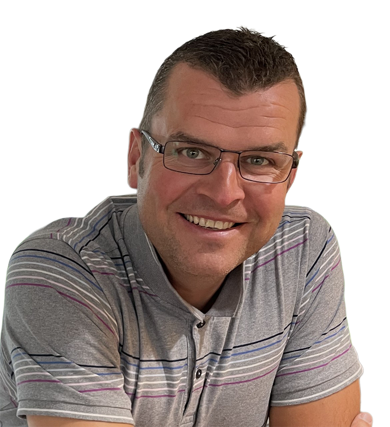 A man with short brown hair and glasses smiles at the camera. He is wearing a grey polo shirt with purple and blue stripes and is seated with his arms crossed on a concrete surface in front of him. | Driveways Plus