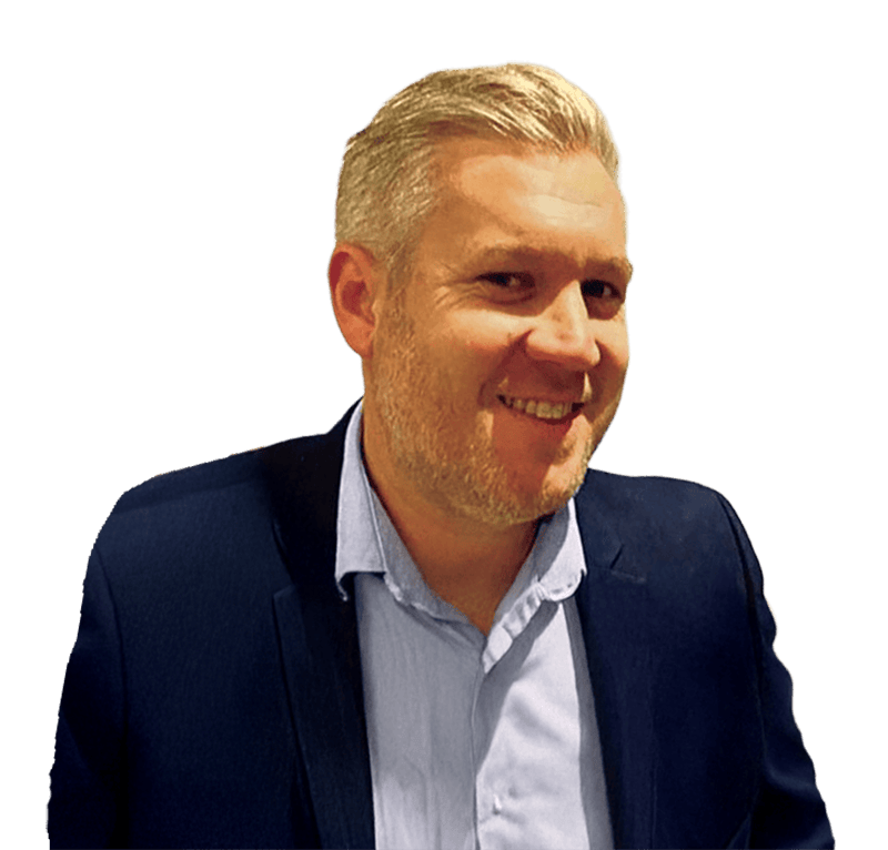 A man with short, light blond hair and a trimmed beard, dressed in a navy suit jacket over a light blue shirt, smiles whilst looking slightly to his left. The background is transparent, resembling the clean lines of fresh block paving. | Driveways Plus