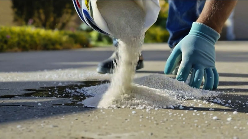 A person wearing blue gloves pours white granules, possibly ice melt or salt, onto a paved surface outdoors—an effective way to remove oil stains from your drive. The background is slightly blurred, showing greenery and sunlight. | Driveways Plus