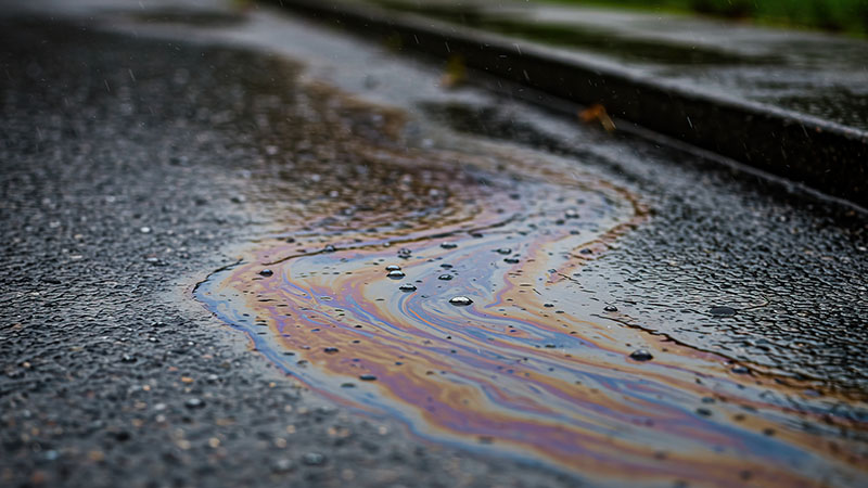 Close-up of a wet asphalt road with a colourful oil slick creating rainbow patterns—reminding you how important it is to remove oil stains from your drive before they spread on rainy days. | Driveways Plus