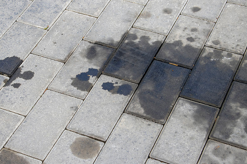Grey stone pavement with several irregular dark stains, likely from water or oil, spread across multiple bricks. The spots vary in size and shape, making it clear why you might want to remove oil stains from your drive. | Driveways Plus