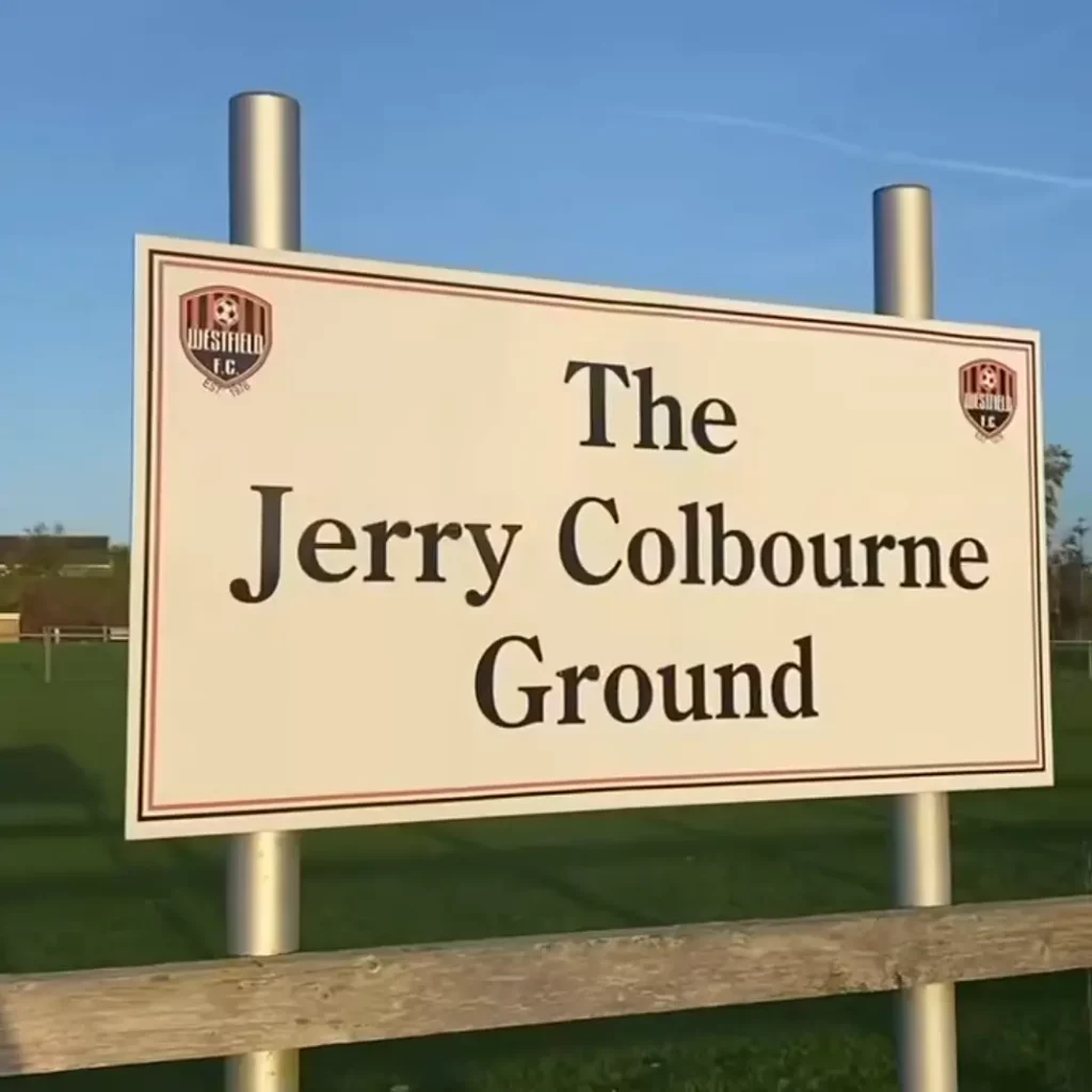 A white sign with black text reads "The Jerry Colbourne Ground," celebrating Westfield FC's 50th anniversary. Two football club logos are in the top corners, and the sign is mounted on metal posts in front of a grassy pitch. | Driveways Plus