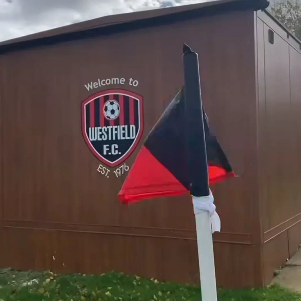 A red and black corner flag stands before a brown building featuring the Westfield FC crest and the words "Welcome to Westfield F.C. Est. 1976," celebrating the football club's 50th anniversary. | Driveways Plus