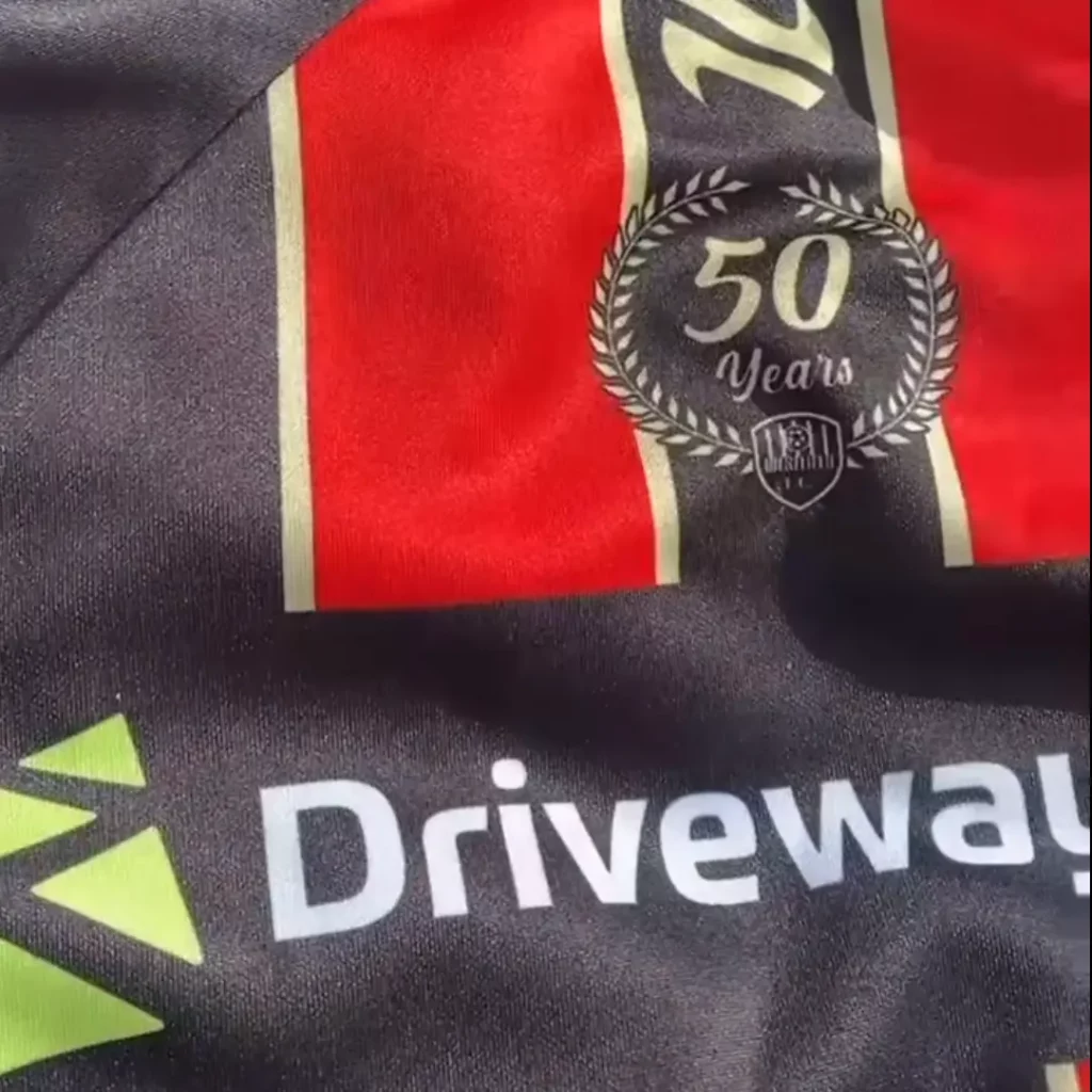 A close-up of a Westfield FC sports kit celebrating its 50th anniversary, featuring a "50 Years" laurel emblem, black and red stripes, and the sponsor logo "Driveway" with a green geometric icon. | Driveways Plus