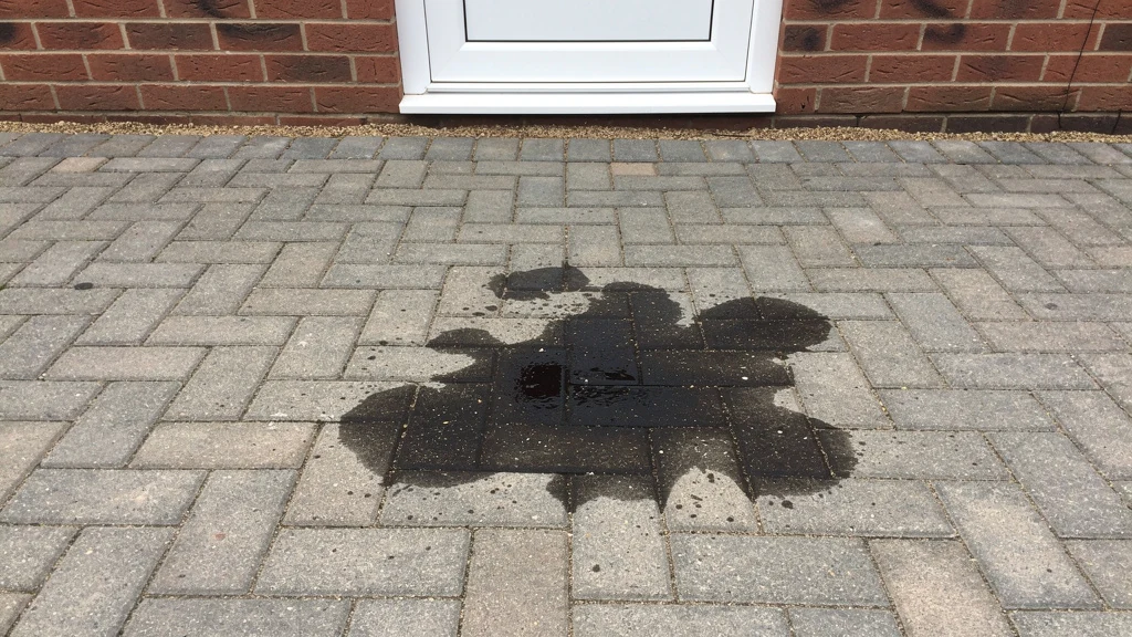 A fresh engine oil stain soaked into a grey block paving driveway laid in a herringbone pattern, beside a red-brick house wall. | Driveways Plus