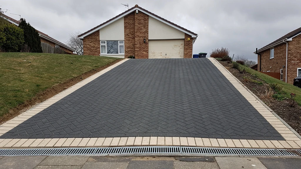 A steep block paving driveway rising to an integral garage, with a full-width channel drain across the foot of the slope at the pavement. | Driveways Plus