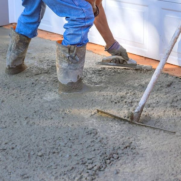 A person in jeans and wellington boots smooths wet concrete with a hand trowel near a garage door in Bristol, while another tool helps level the surface. | Driveways Plus